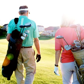 Two people walk together across a golf course while carrying their golf bags.