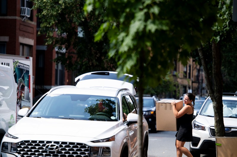 A person packs a car trunk outside on a sunny day in a Boston neighborhood.