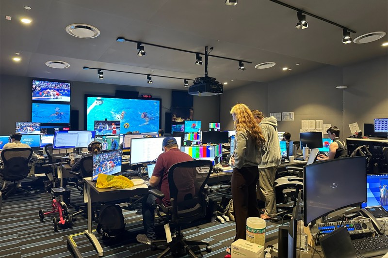 People sitting in a room full of monitors displaying Olympics coverage.