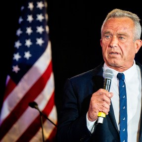 RFK Jr speaking into a microphone in front of the United States flag.