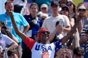 Snoop Dogg wearing a USA shirt at a women's beach volleyball game with his arms raised in the air.