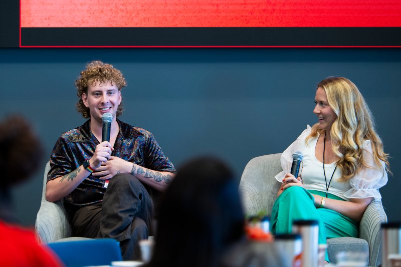 Cam Sleeper sitting on stage speaking into a micrphone while Emily Schario sits next to him watching him speak. 