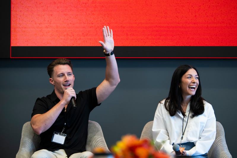 Kevin Cooney speaking into a microphone with one hand raised in the air and Ashley DeMato-Cooney sitting next to him laughing.