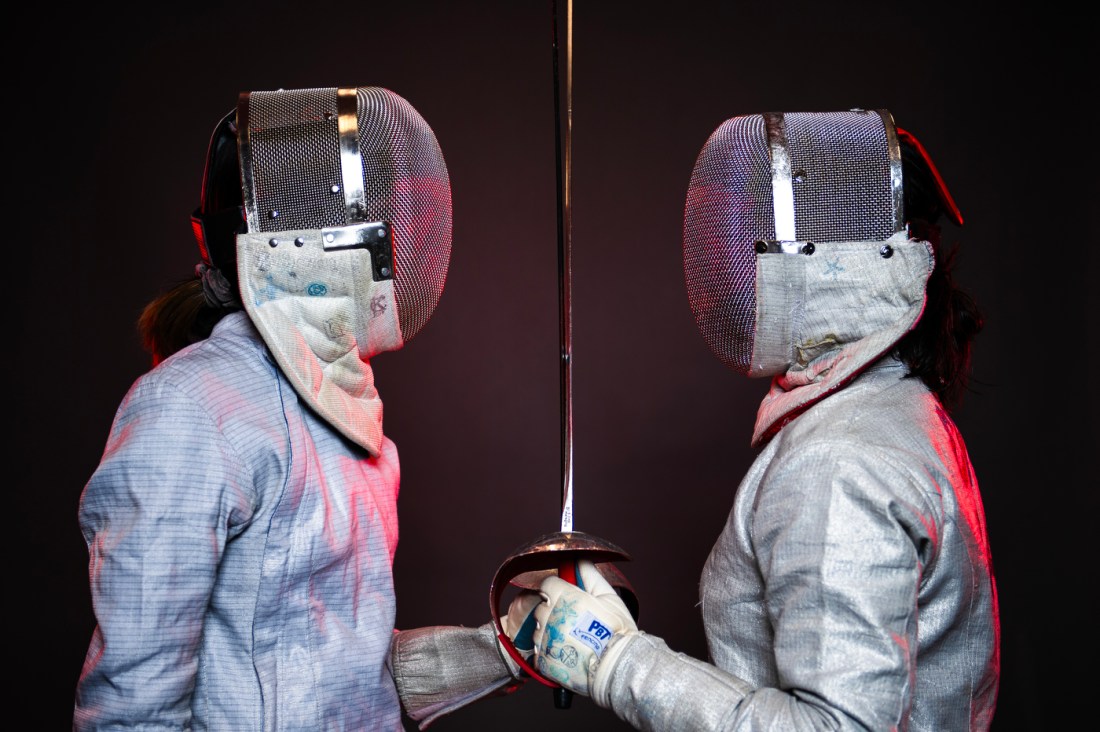 Two people in fencing gear facing each other while holding their swords up in perfect alignment in between them. 