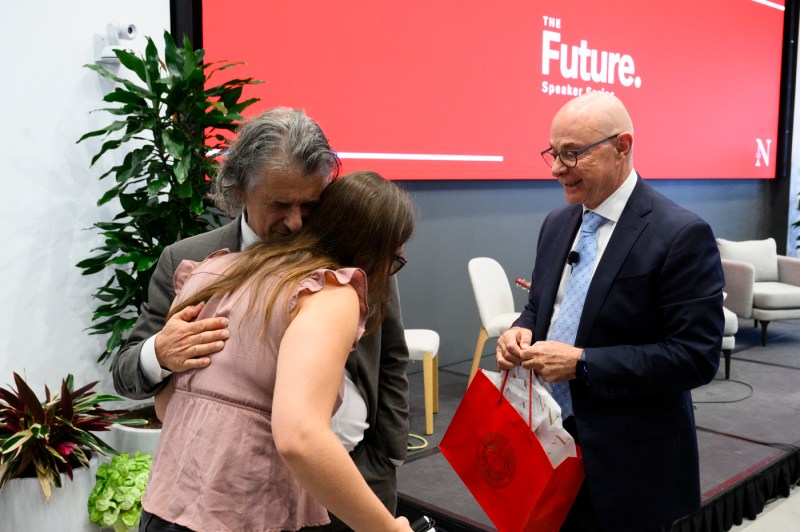 Daniel Levitin hugging and greeting another person while President Aoun stands next to them, holding a Northeastern branded red gift bag.