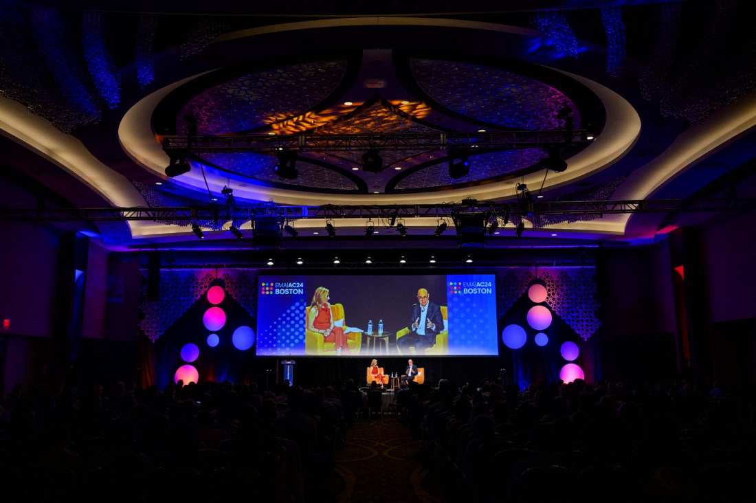 Aoun and Claire Goldsmith sit in orange chairs on stage at the Sheraton Boston Hotel, with a large screen behind them livestreaming the event to an audience in the back of the venue.