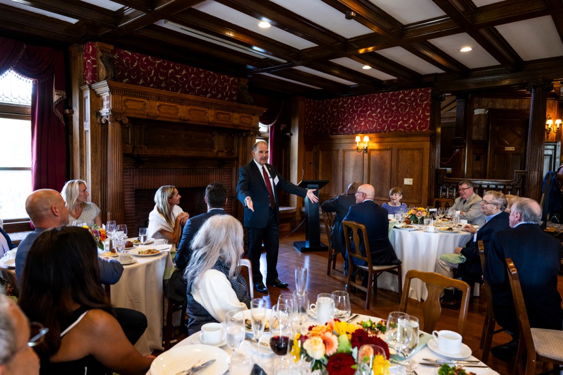 A speaker standing up in a room full of people and giving a speech at a luncheon.