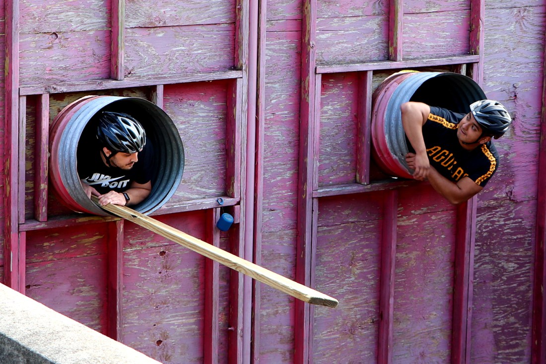 Two students wearing bicycle helmets crawling out of pipes. 