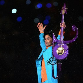 Prince wearing a blue jacket holding a purple guitar up in the air while performing at a Super Bowl half-time show.