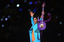 Prince wearing a blue jacket holding a purple guitar up in the air while performing at a Super Bowl half-time show.