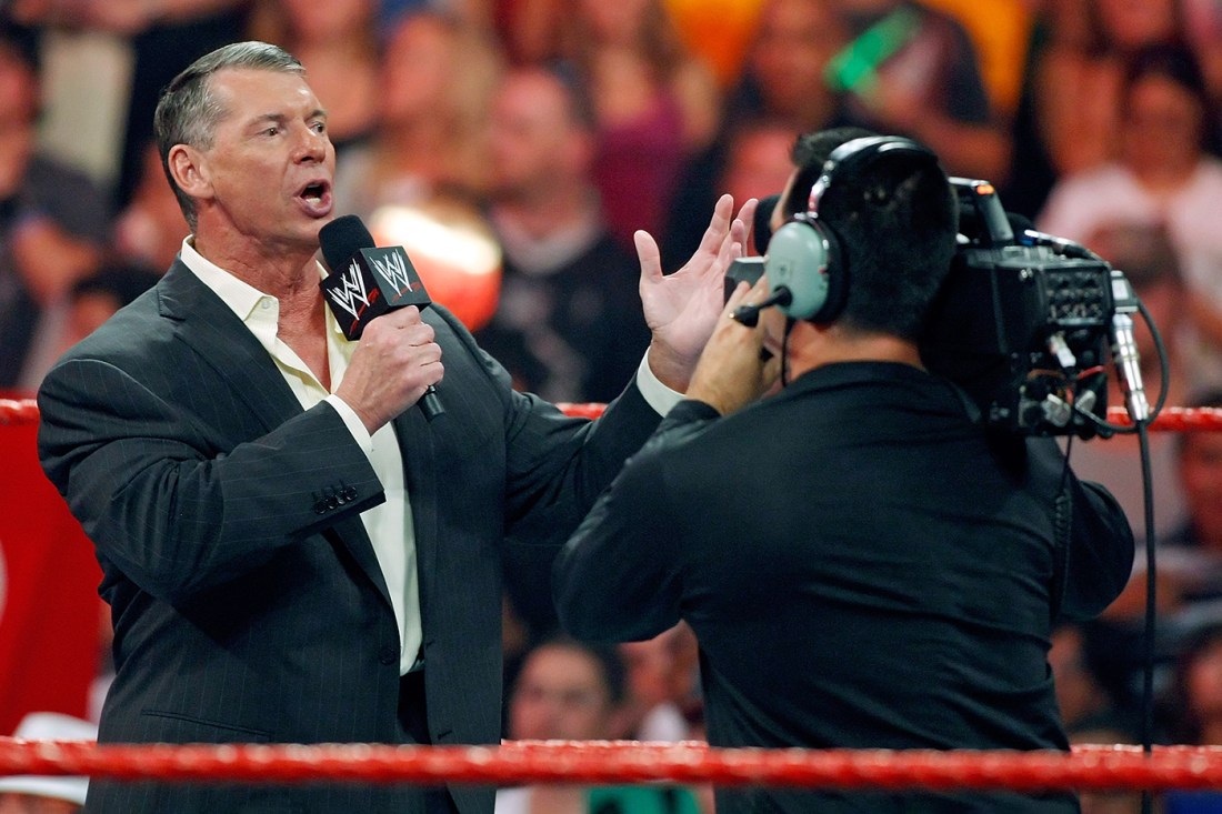 Vince McMahon standing in the ring during a WWE match speaking into a microphone in front of a TV broadcast camera.