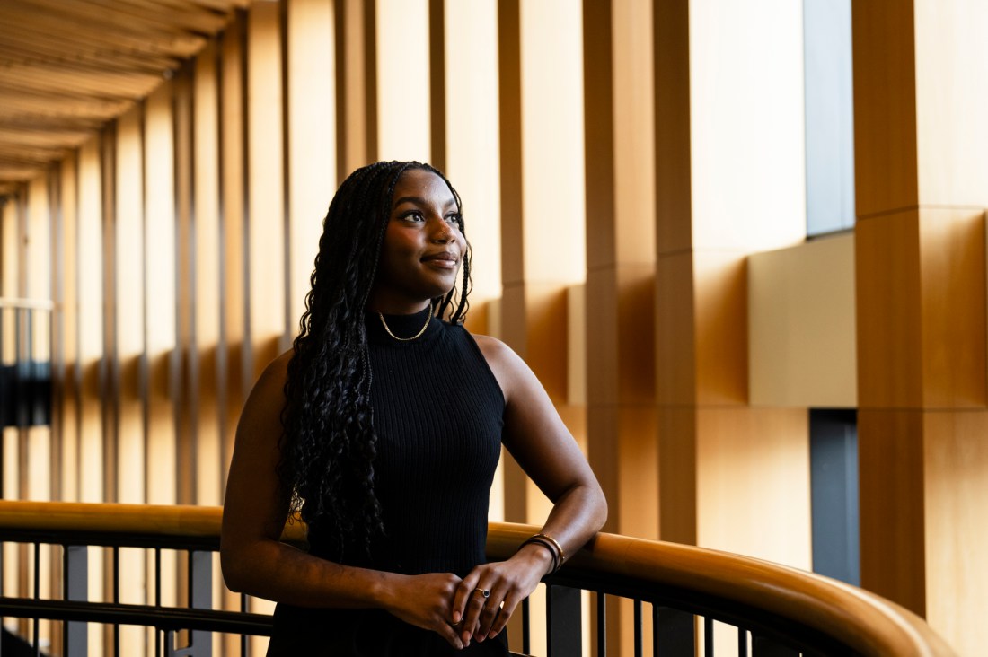 A student standing near large windows in a bright hallway, smiling and posing for a portrait.