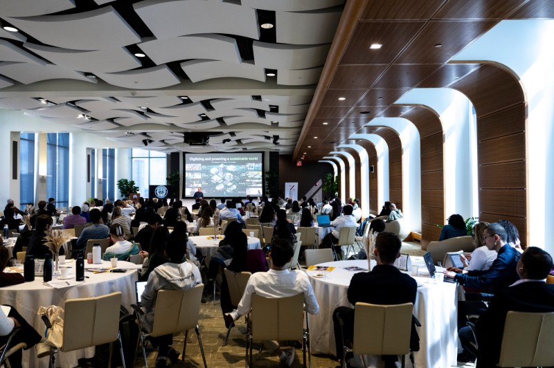 A large audience sitting at round tables listens to a person deliver a presentation.