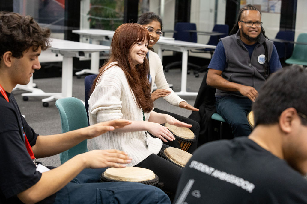 A group of people sat in a circle, smiling and playing hand drums together.