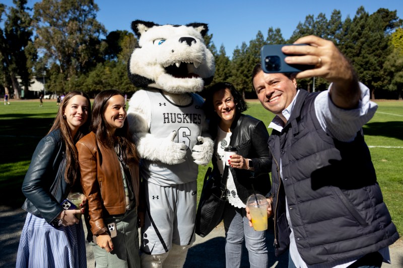 A group of people pose for a selfie outdoors with a husky mascot.