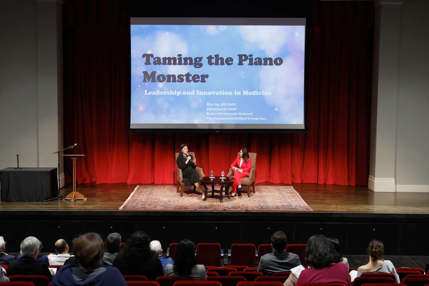 Dr. Rita Ng sitting next to another woman on stage under a screen that displays the title 'Taming the Piano Monster: Leadership and Innovation in Medicine'.