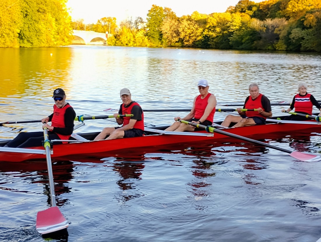 Five 80-year-old rowers in a boat in the Charles River wearing red shirts.