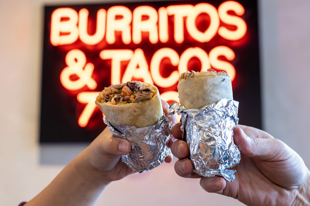 Two people's hands holding up wrapped burritos in front of a neon sign that says 'Burritos & Tacos'