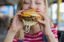 A girl with bright pink painted fingernails biting into a fast food burger.