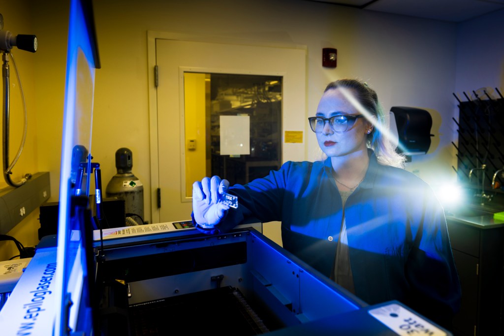 A person in a lab coat works with scientific equipment in a laboratory setting, holding a small object under bright light.