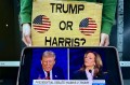 A screen showing a broadcast of the presidential debate. Behind it a person holds a yellow sign that says 'Trump or Harris?' on it with two circle American flags.