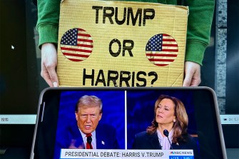 A screen showing a broadcast of the presidential debate. Behind it a person holds a yellow sign that says 'Trump or Harris?' on it with two circle American flags.