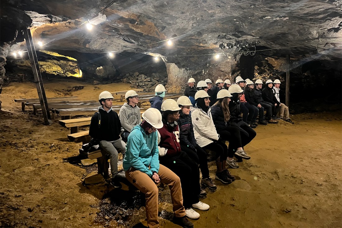 A group of students wearing white hard hats in an underground cavern, sitting on benches. 