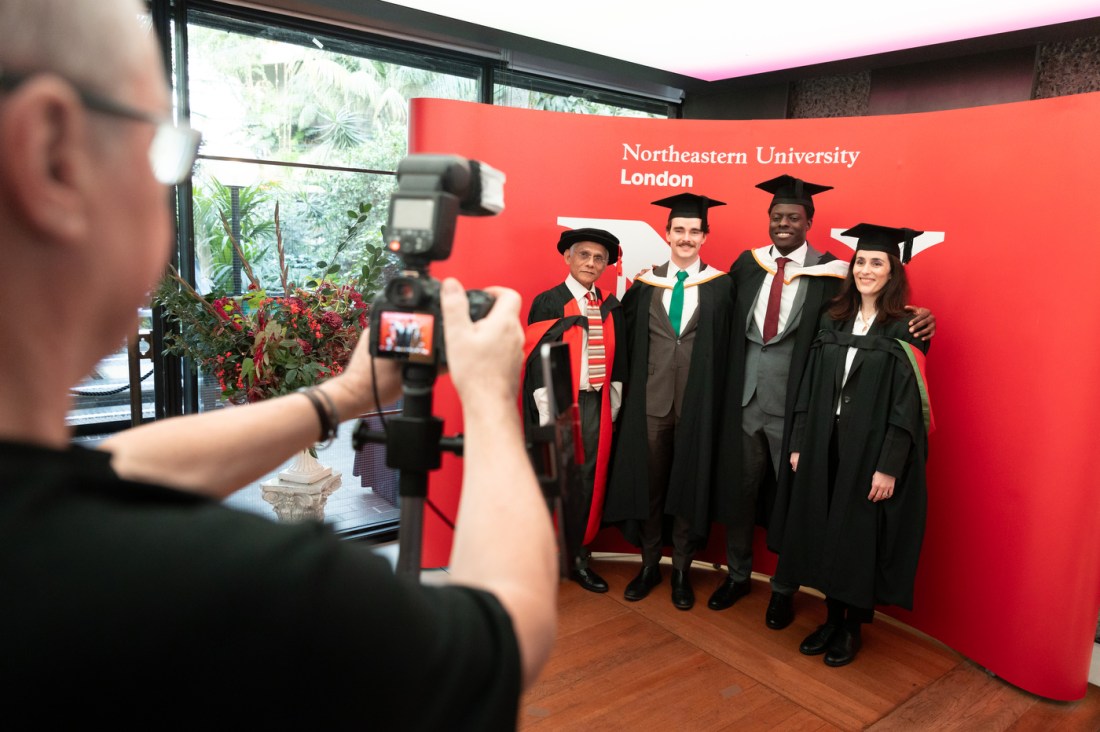 Four graduates posing in front of a red backdrop in front of a professional photographer.