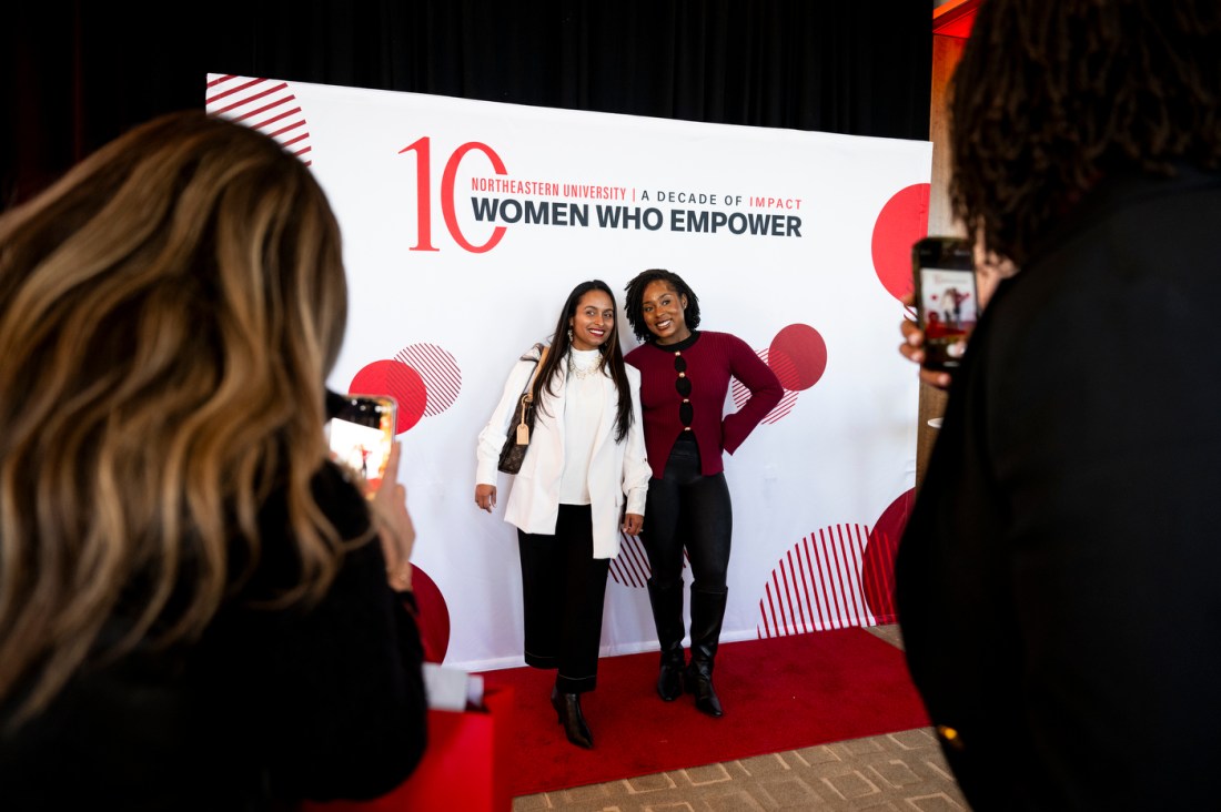 Two people posing in front of a Women Who Empower backdrop. 