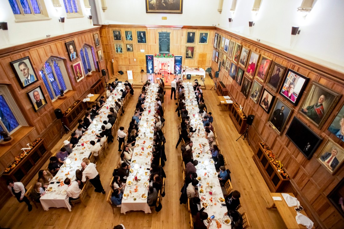 A large wood-paneled dining hall filled with students seated at long tables, with Joseph E. Aoun speaking at the front.