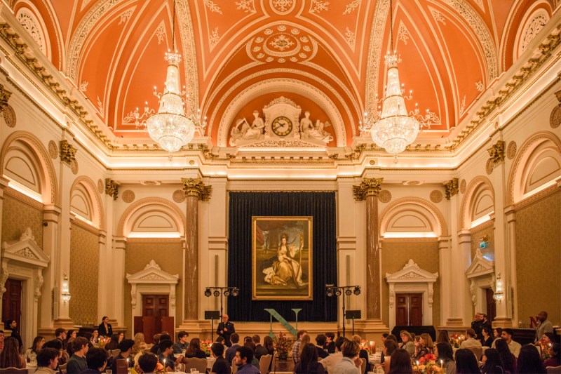 An ornate dining hall with chandeliers and high ceilings filled with students seated at long tables.