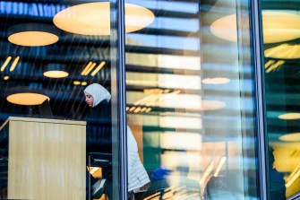 A student studies at a desk inside a modern building with large windows and bright overhead lights.