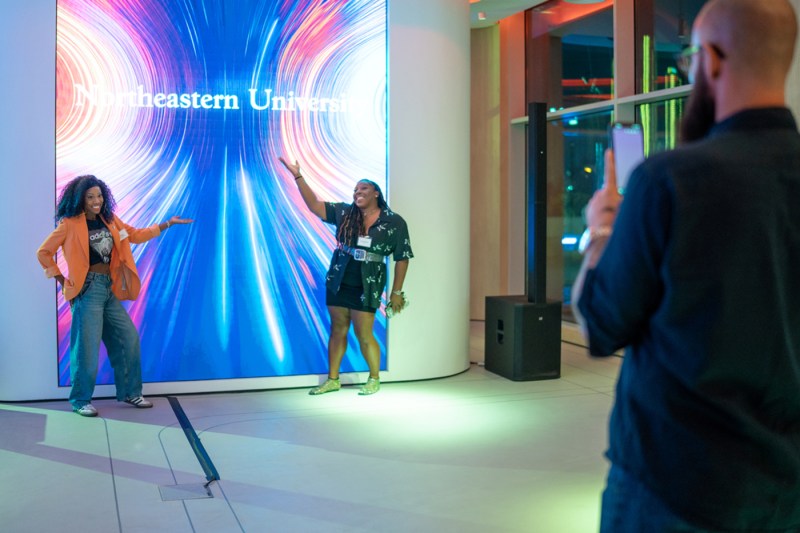 Two people posing with a colorful LED Northeastern sign.