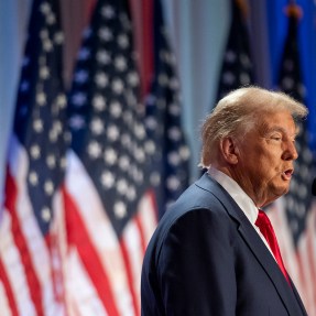 Donald Trump speaking on a stage in front of a row of US flags.