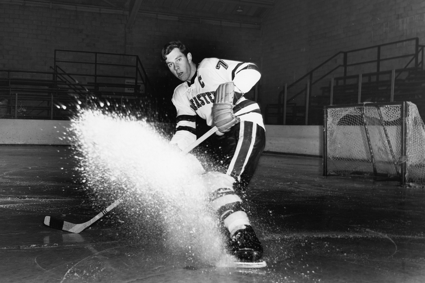 A black and white photo of David Poile wearing a Northeastern hockey jersey and sliding to a stop on the ice. 