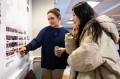 Sasha Hearn wearing a sweater pointing at a project displayed on the wall and explaining it to Feifei Shen, who is leaning in to get a closer look.