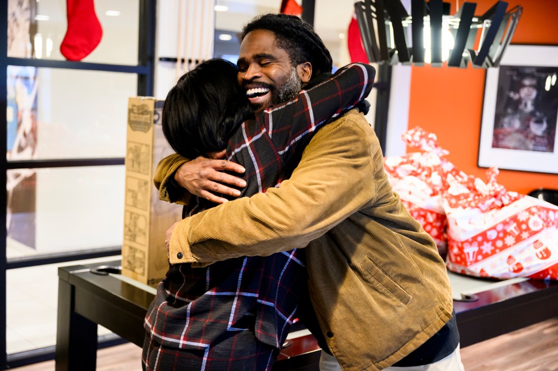 A man in a light brown jacket smiles as he hugs a woman with dark hair, wearing a checkered sweater.