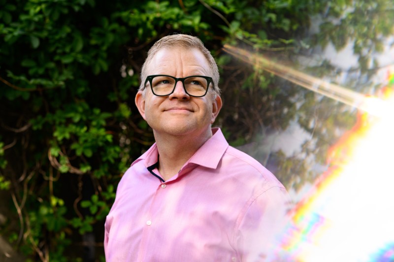 William Sharp, wearing glasses and a pink polo shirt, smiles while standing outdoors against a backdrop of lush green foliage, with an artistic rainbow prism light effect overlaying part of the image.