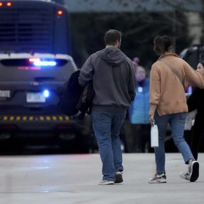 Three people walking outside of a school in Wisconsin by a police car with it's lights on.