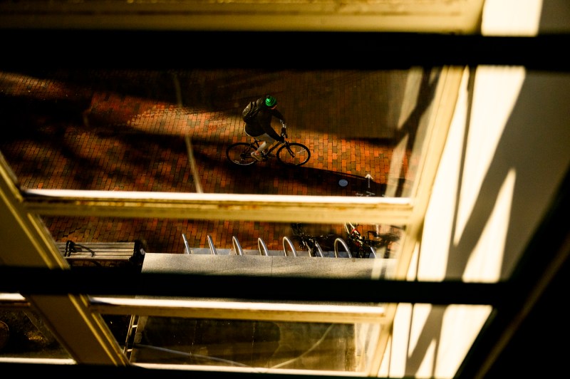 A student wearing a green helmet rides a bike on a brick pathway past a university campus framed by window panes and shadows.