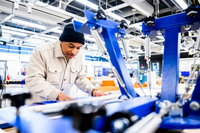 A person works on a project in a modern makerspace with industrial equipment and bright lighting.