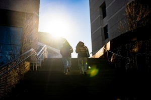 Two individuals walk up an outdoor staircase with sunlight in the background on a clear day.