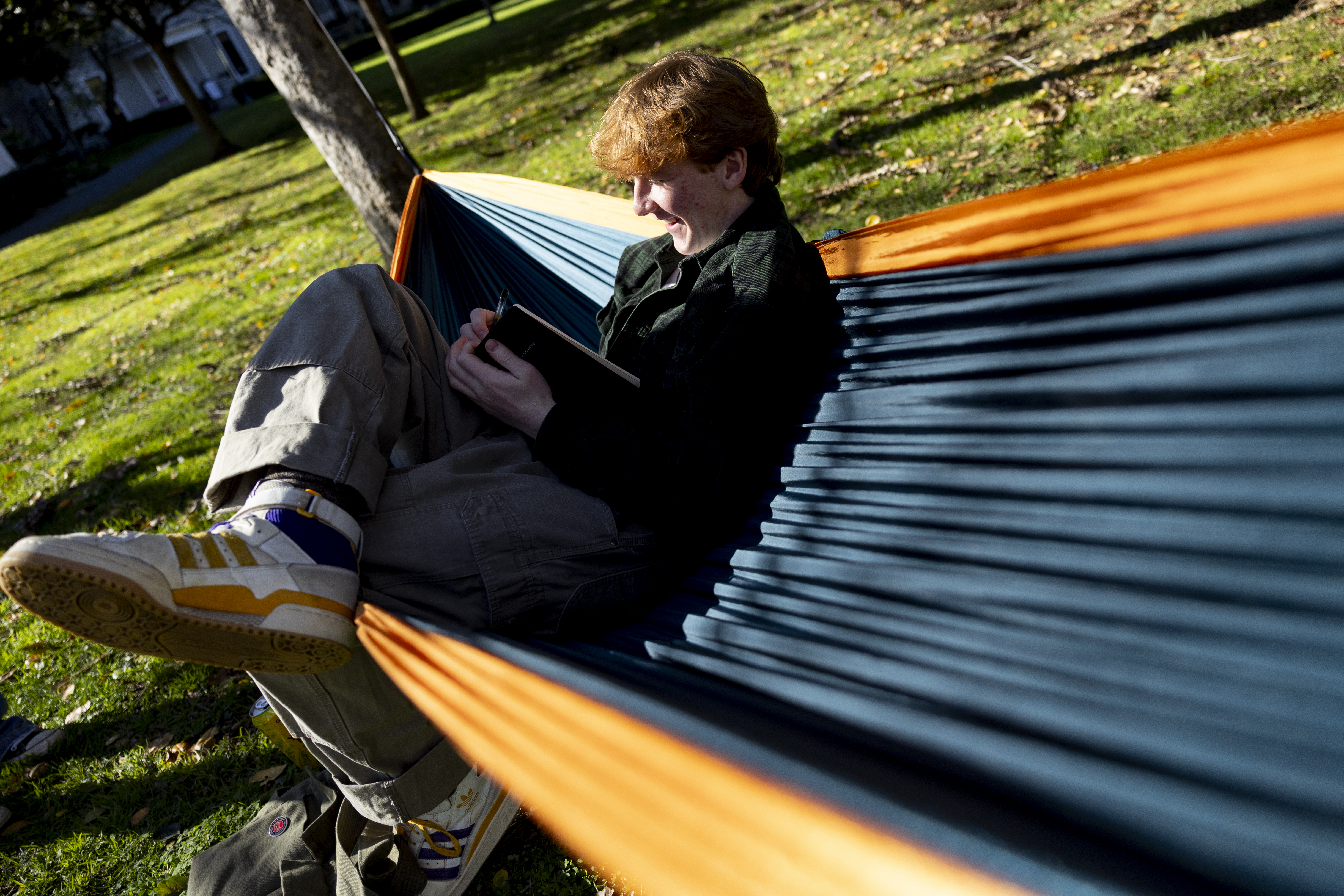 A student sits in a yellow and blue hammock strung between. two trees reading a book with one leg crossed over the other.