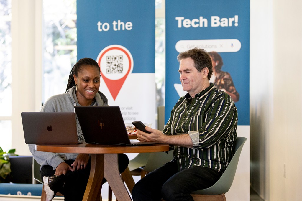 A person assists another at a table with laptops, both engaged in conversation. A backdrop with signs suggesting a tech-related setting.