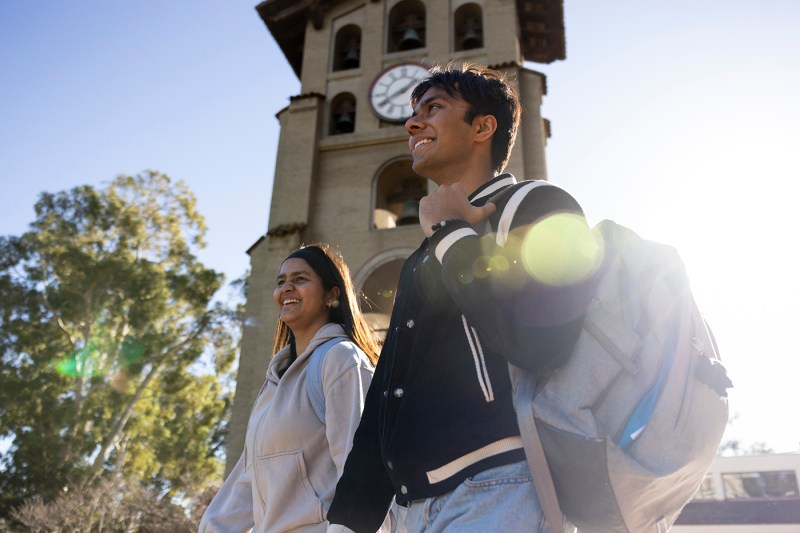 Two students walk outdoors on a sunny day near a clock tower.