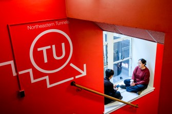 A bright red wall with text and symbols, featuring two people sitting and talking near a window in an indoor setting.