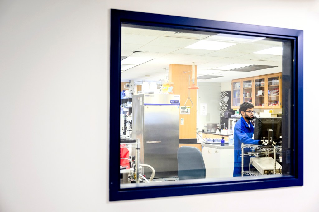 A view through a window into a laboratory where someone works, surrounded by equipment and supplies.