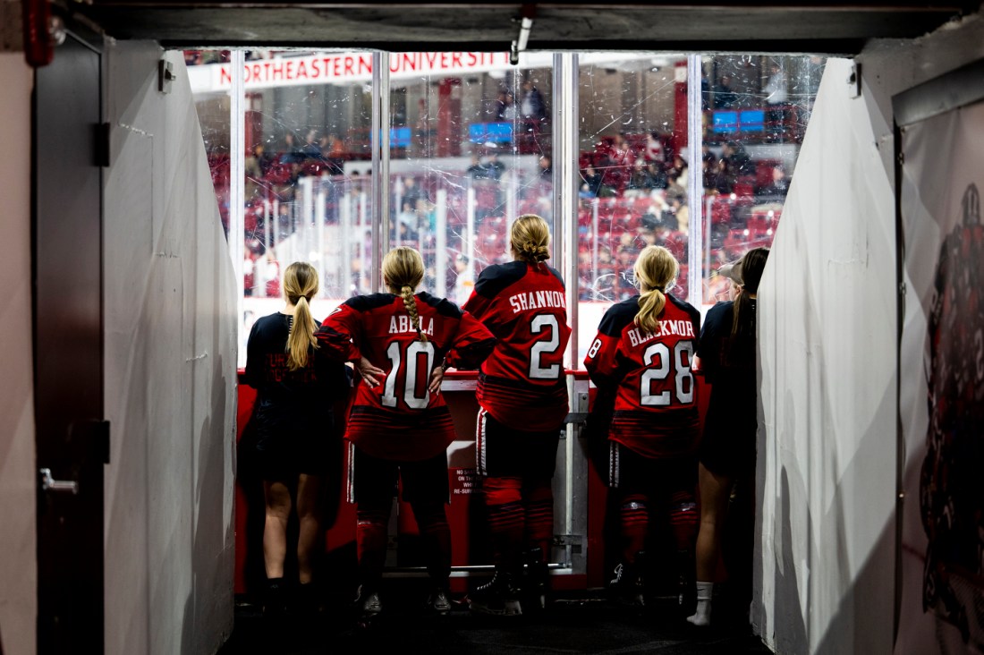 Northeastern women’s hockey players walking through a brightly lit tunnel toward the ice, seen from behind.