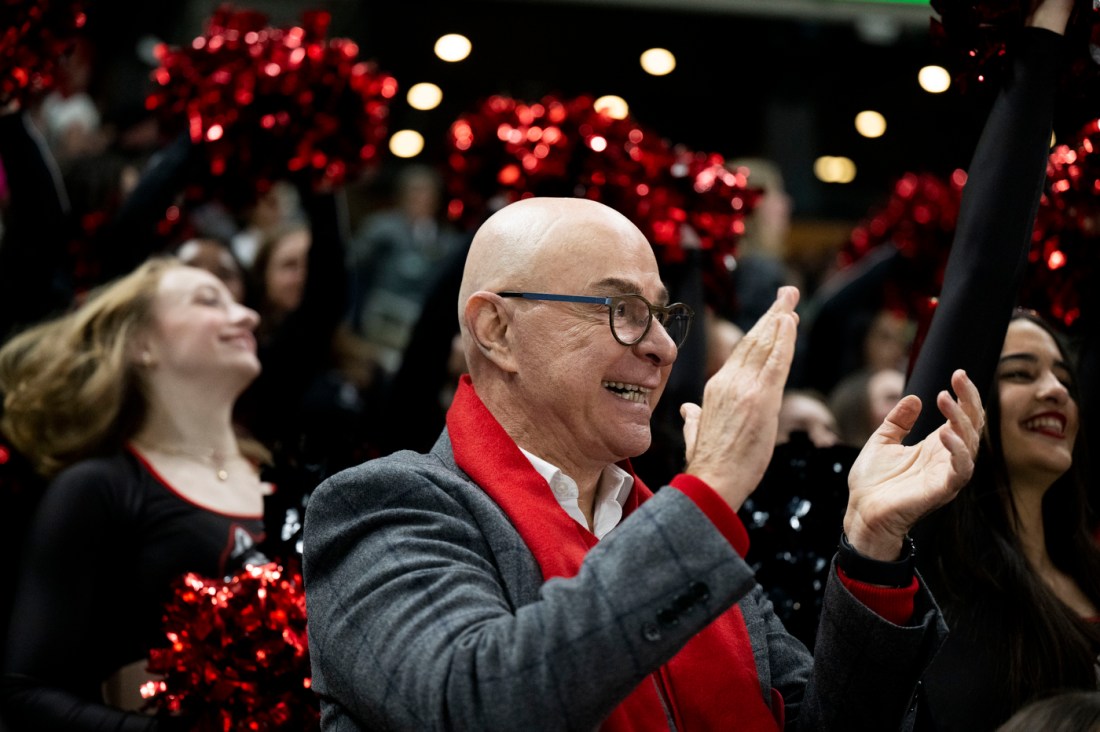 Northeastern President Aoun wearing a red scarf and a grey blazer claps and smiles while cheering from the stands.