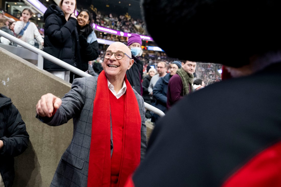 Northeastern President Aoun enters the arena smiling wearing a red scarf, a red sweater, and a grey blazer over top.